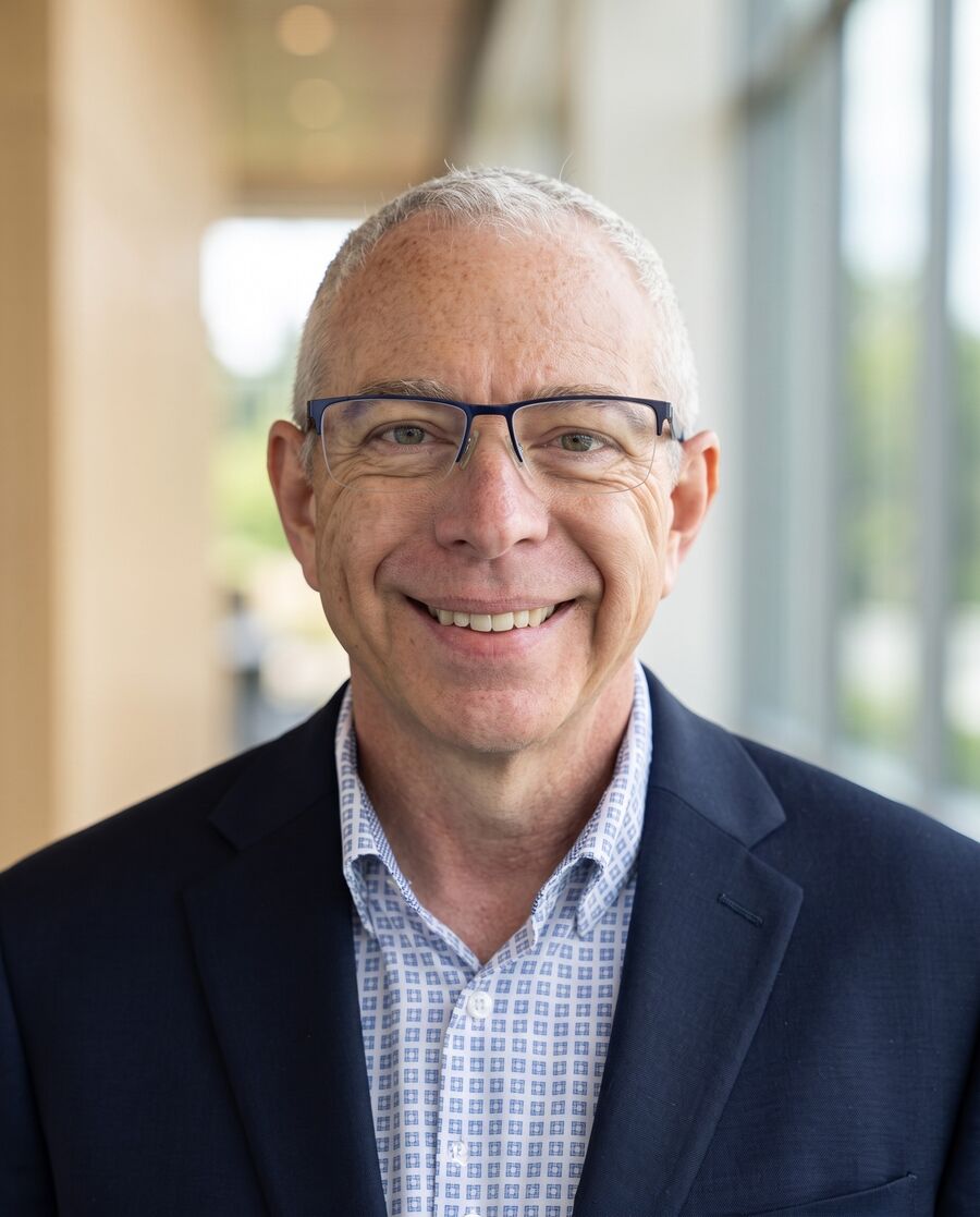 Brian Olson smiling, in a navy blazer and patterned blue shirt, with a warm corridor and windows behind him.