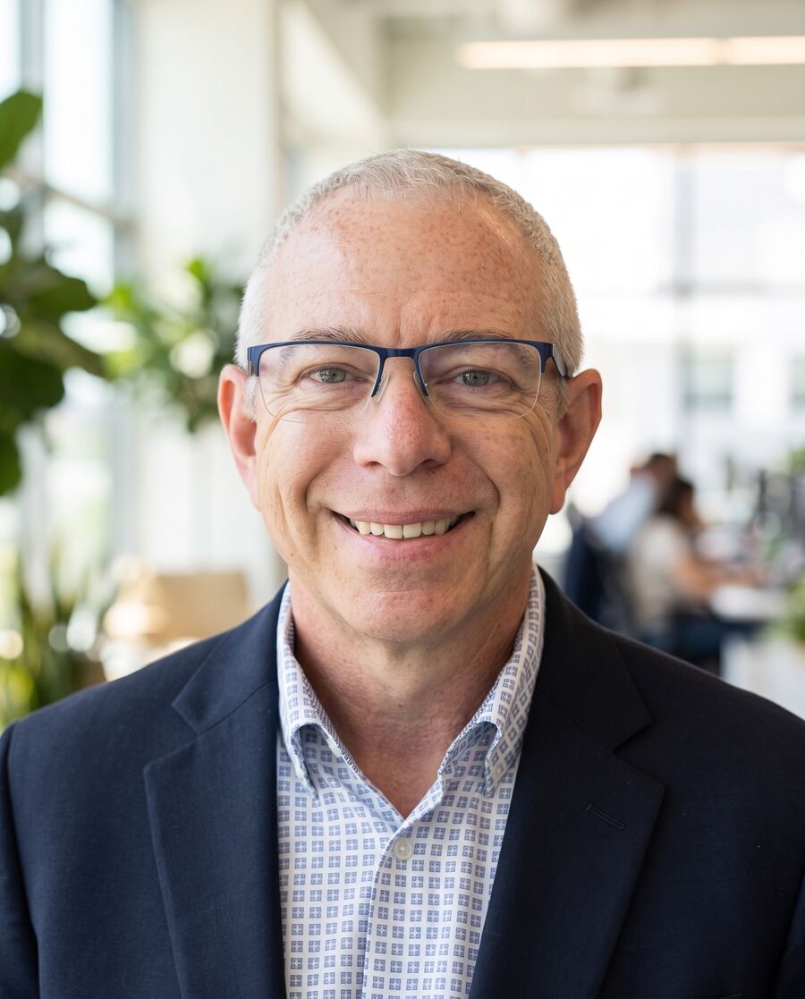 Brian Olson smiling in a bright open workspace, plant to one side and people softly blurred behind him.