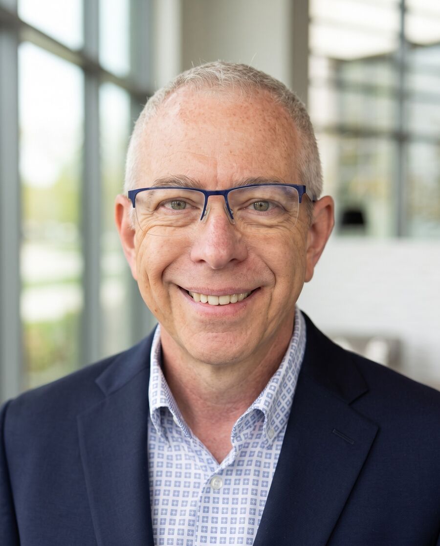 Brian Olson smiling in front of floor-to-ceiling windows with greenery visible outside.