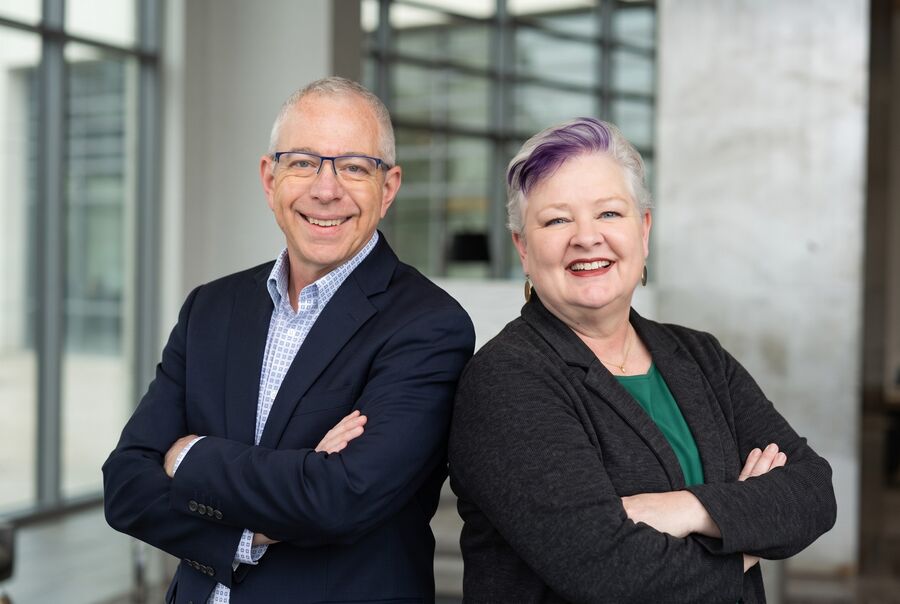 Brian and Robin Joy Olson standing back to back with arms crossed, smiling, in a glass-walled atrium.