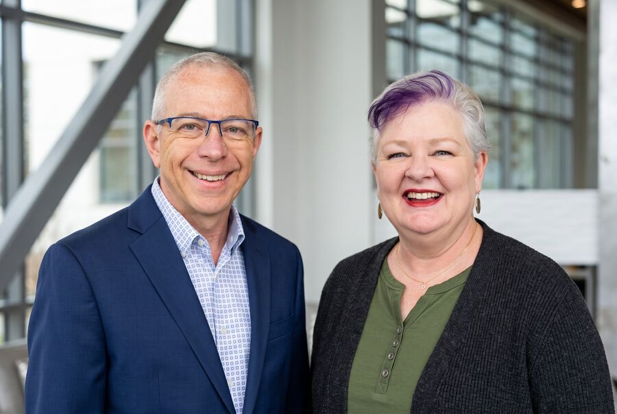 Brian and Robin Joy Olson smiling in a building atrium, Robin in an olive henley and charcoal cardigan.