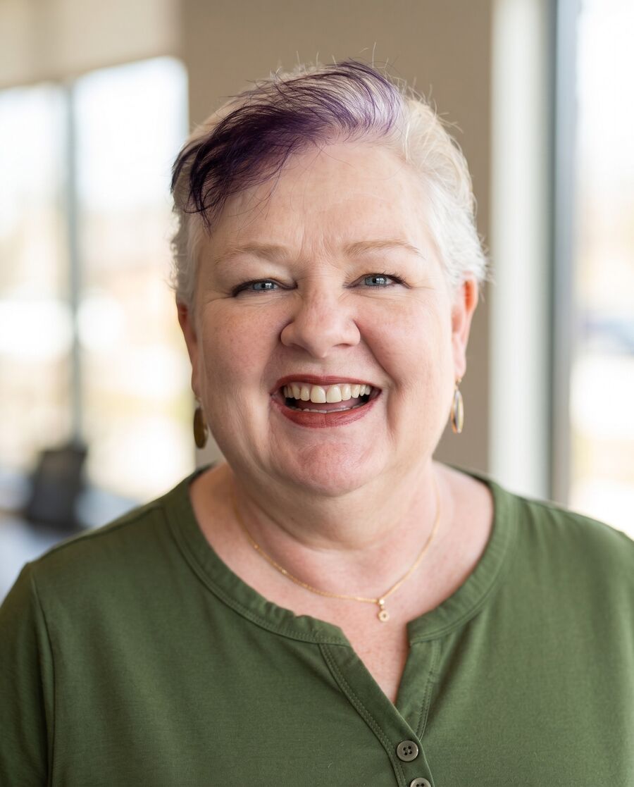 Robin Joy Olson laughing, in an olive green henley, with a warm wall and window light behind her.